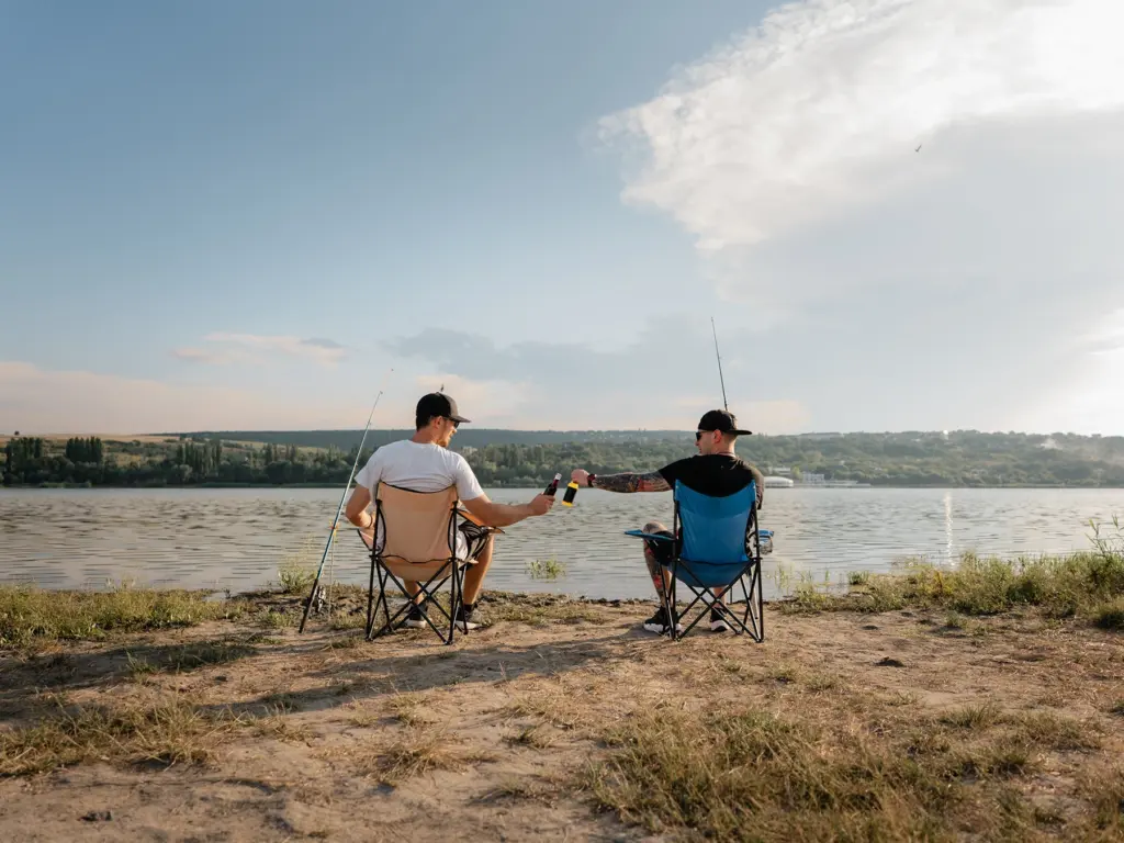 Two men fish from folding chairs on a riverbank while toasting drinks, an idea for where to travel in august.