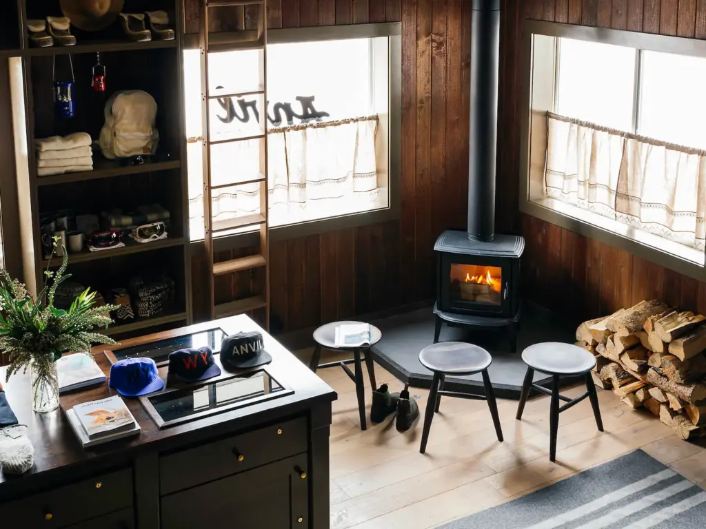 Wood-paneled cabin interior with a cast iron wood stove, small stools, stacked firewood, and shelves holding hats and goggles.
