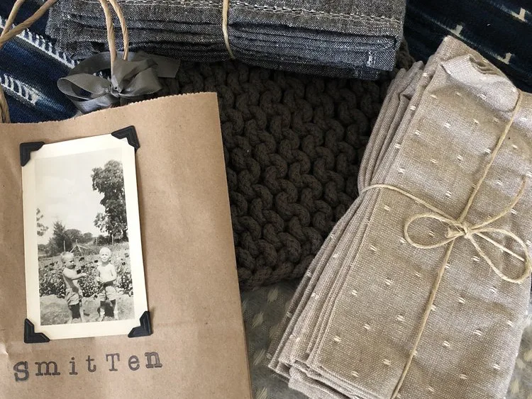 Kraft paper bag labeled Smitten with vintage photo beside folded textiles and chunky knit fabric at a shop in Fredericksburg TX.