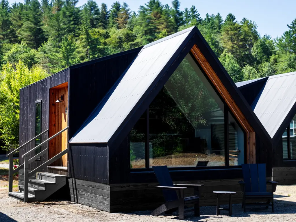 Modern black A-frame cabin with a large front window and a small wooden porch nestled near a pine forest.