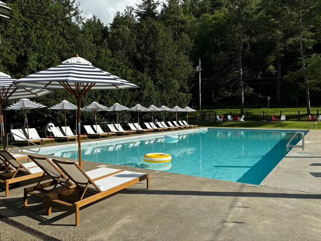 Lounge chairs and black-and-white striped umbrellas surround a large outdoor swimming pool on a sunny day.