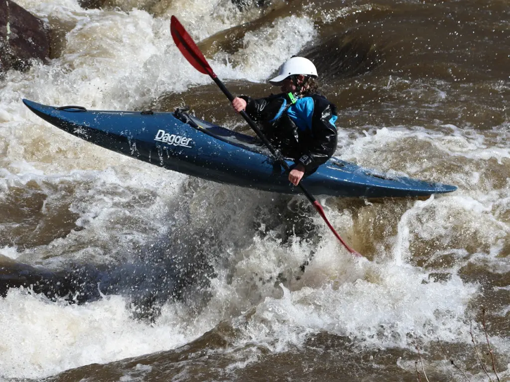 Kayaker paddling a blue kayak through rough whitewater rapids, an adventurous choice for where to travel in august.