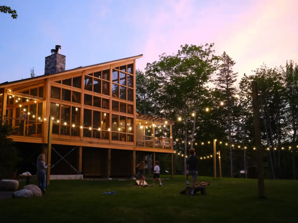 String-lit glass and wood cabin at dusk, showcasing design-forward nature retreats where guests gather on a glowing lawn.