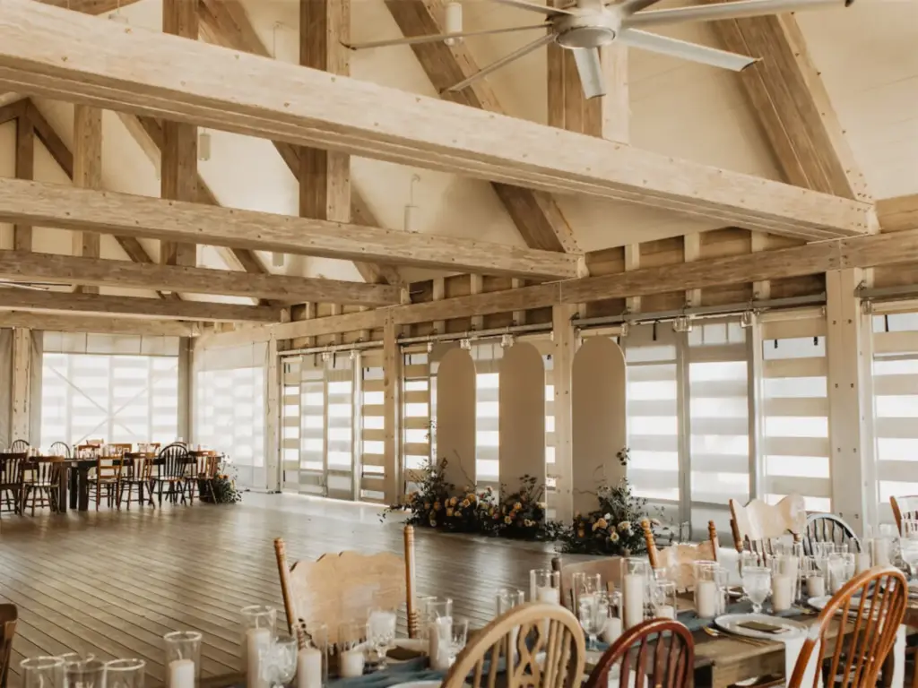 Rustic barn wedding reception hall with exposed wood beam ceiling, long dining tables, and floral arrangements on wood floors.