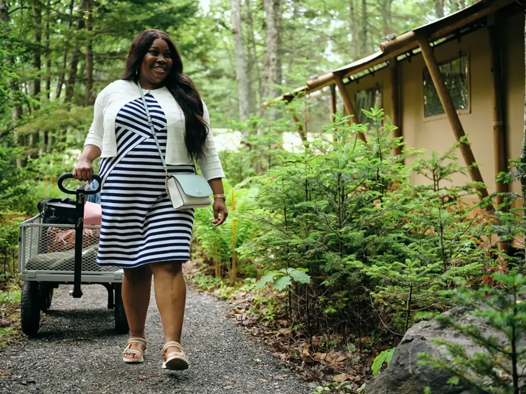 Woman in striped dress pulling a utility wagon on a wooded gravel path beside a tent cabin.