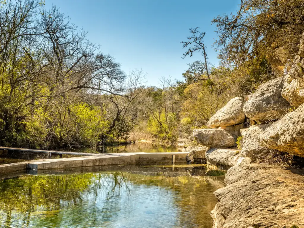 A scenic swimming hole on one of the best road trips in Texas, with limestone rocks and green water under a clear blue sky.