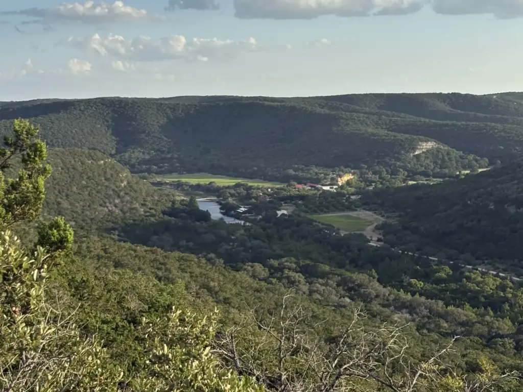 Aerial view of one of the best road trips in Texas: a wooded Hill Country valley with a river, green fields, and rolling hills.