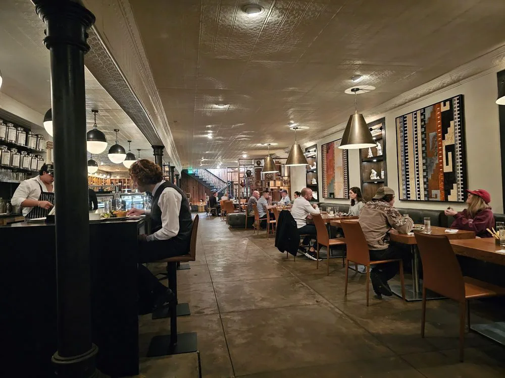 Dimly lit restaurant dining room with exposed ceiling tiles, pendant lights, leather chairs, and patrons eating along a long open floor plan.