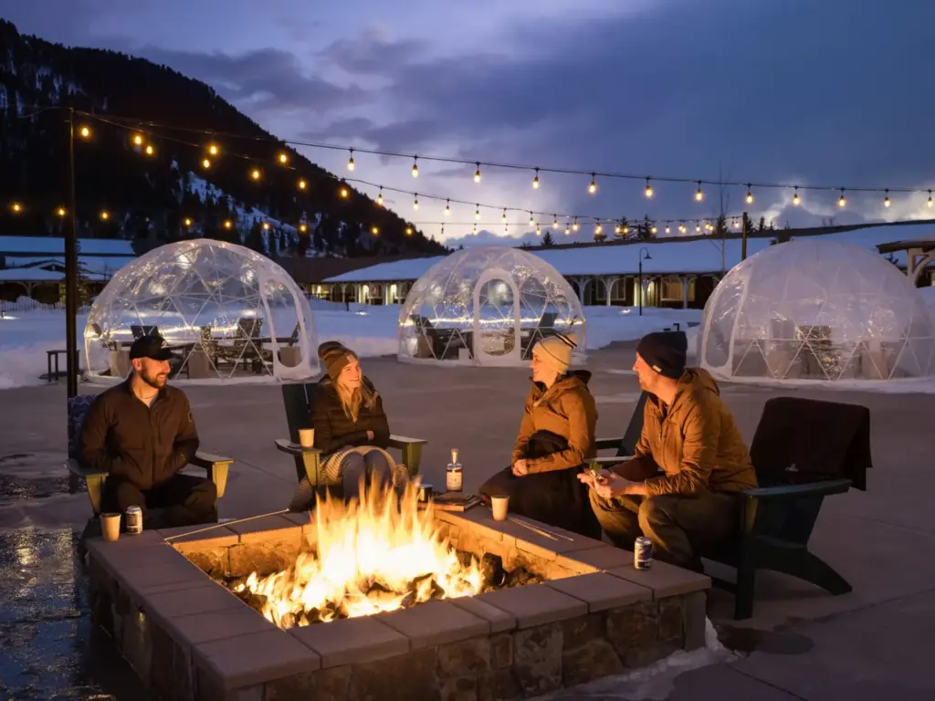 Four colleagues warm up by a fire pit at a nature immersive corporate offsite, with glowing igloo domes and snowy mountains at dusk.
