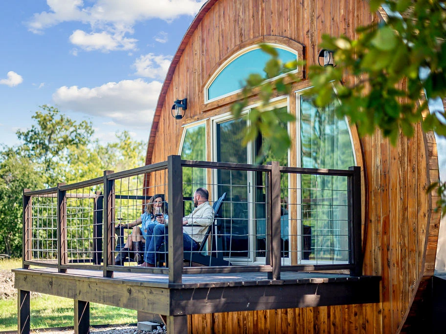 A couple relaxes on the deck of a unique wooden barrel-shaped cabin surrounded by nature.