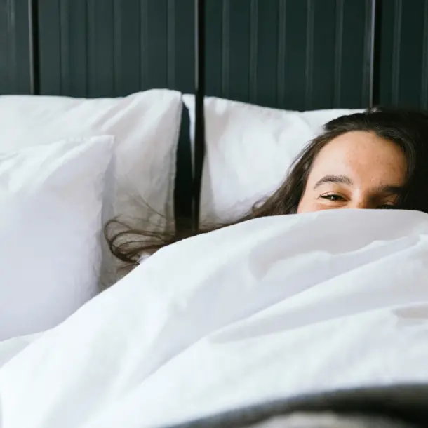 A woman with long dark hair peeks over white bedding with just her eyes visible, lying in a bed with white pillows.