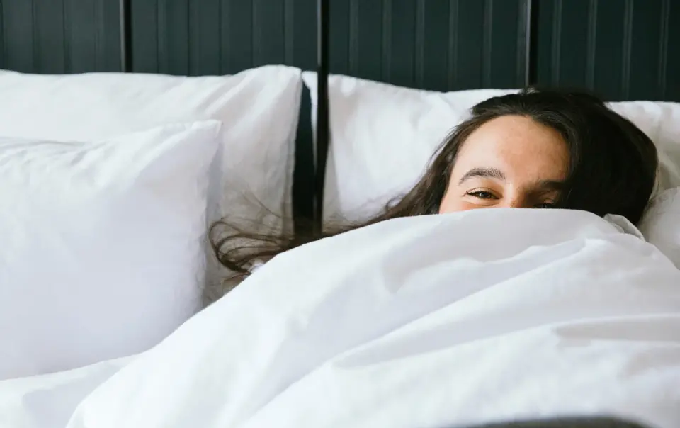 A woman with long dark hair peeks over white bedding with just her eyes visible, lying in a bed with white pillows.