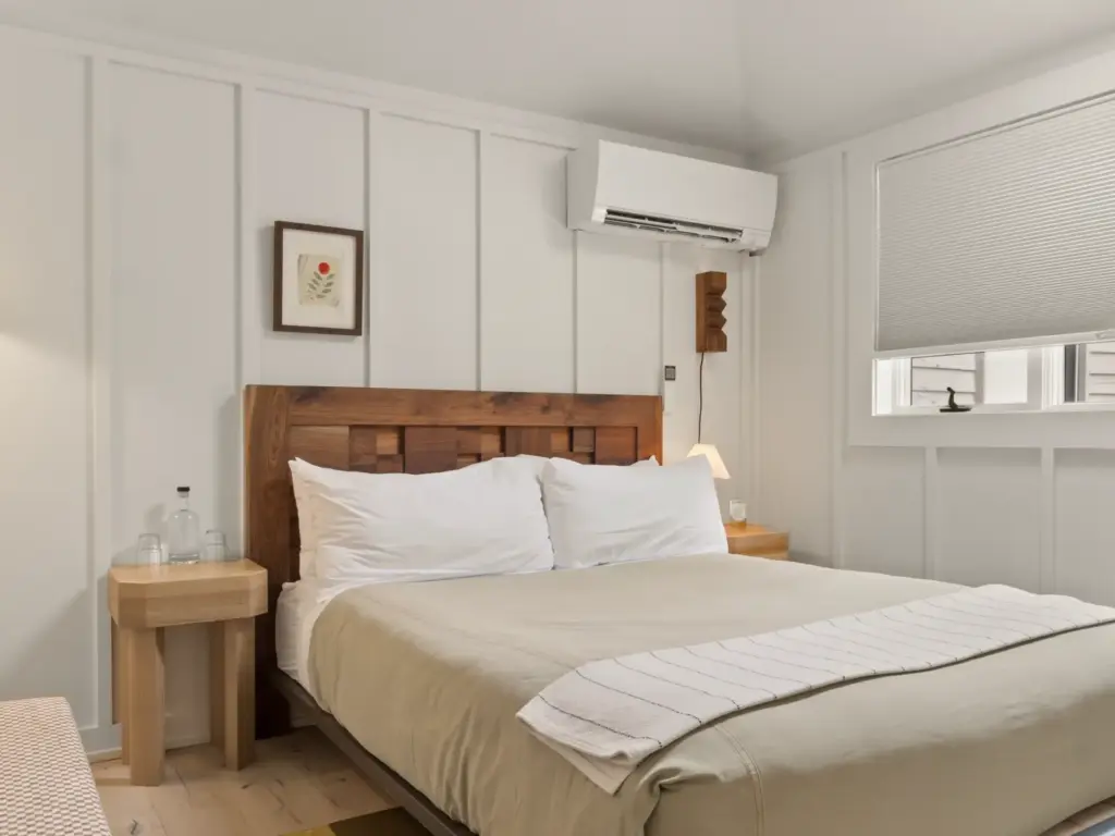 Queen bed with wood headboard, white pillows, and neutral linen duvet in a white paneled bedroom with mini-split AC unit.