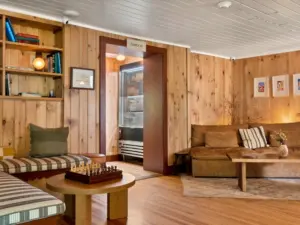 Warm wood-paneled lounge with a leather sofa, chess set on a round table, bookshelf, and a lit refrigerator visible through a check-in doorway.