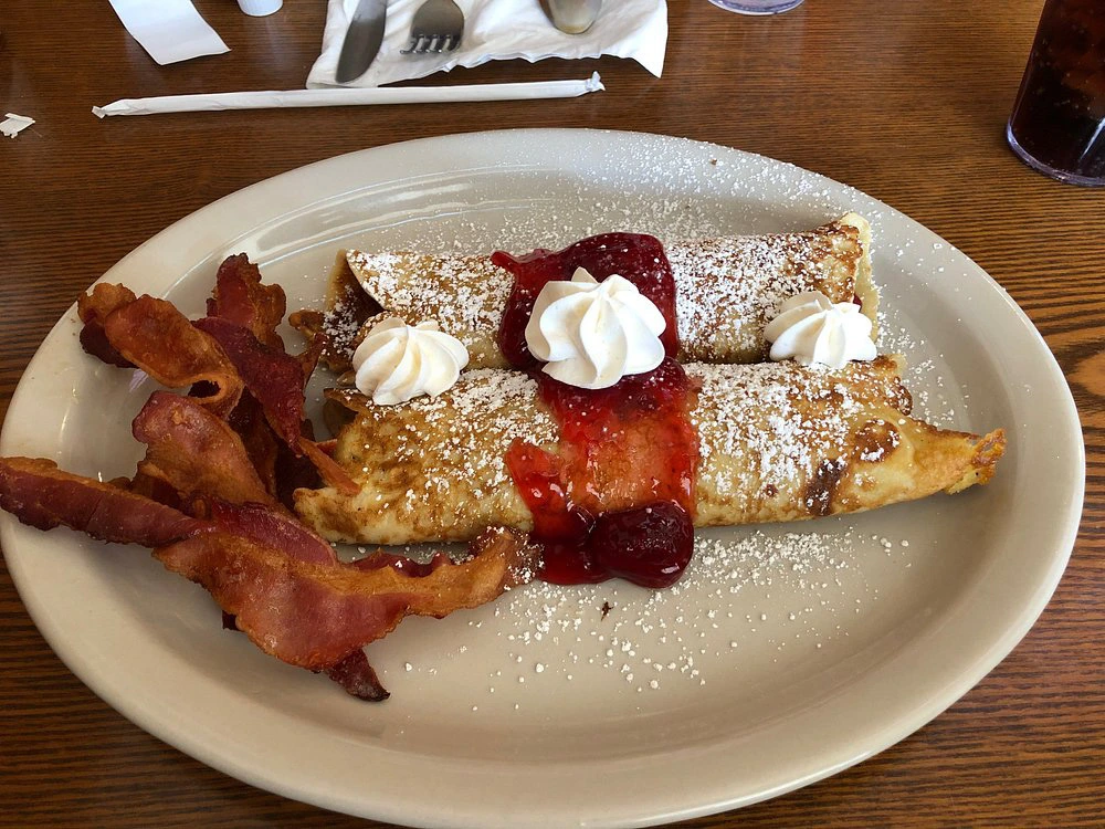 Two strawberry-filled crepes topped with whipped cream, served with a side of crispy bacon.