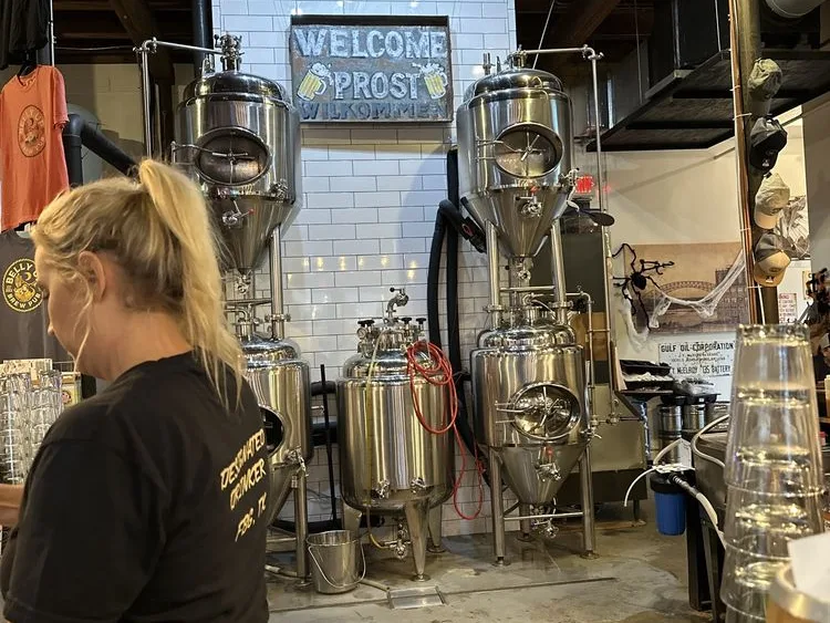 Bartender near brewing tanks in one of the breweries in Fredericksburg Texas, beside a Welcome Prost Wilkommen sign.