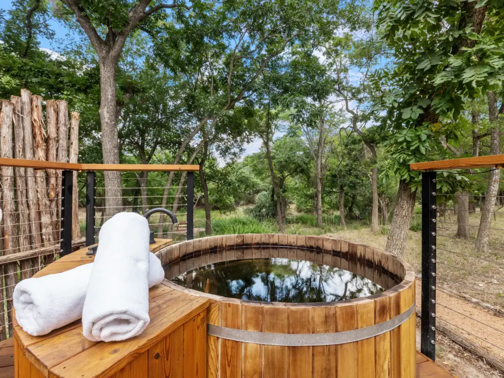 Wooden barrel hot tub on a deck surrounded by trees, with two rolled white towels resting on the rim.