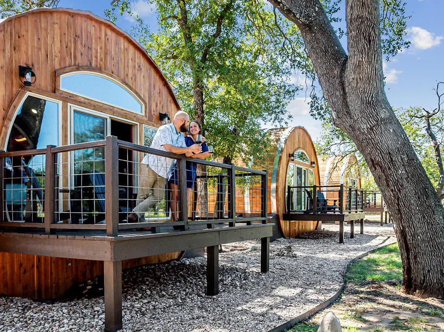 Couple relaxing on the deck of a curved wood barrel-style cabin surrounded by oak trees and gravel paths.
